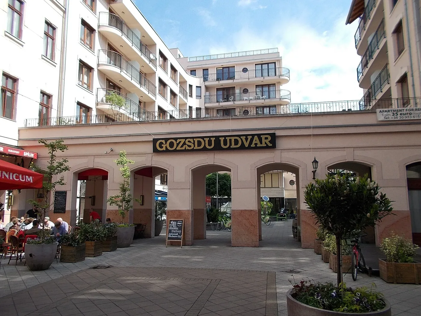 A courtyard inside Gozsdu Udvar in daylight: a five-arched arcade under a black and gold GOZSDU UDVAR sign, terrace tables and a red parasol on the left, planters and young trees across the paving, and a modern balconied apartment block rising above the arcade under open sky