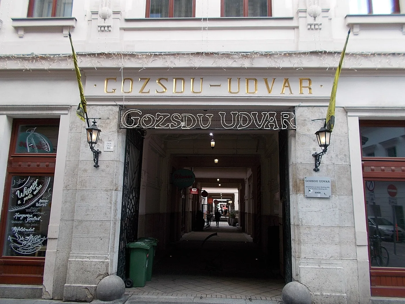 The Dob utca 16 entrance to Gozsdu Udvar: a stone archway with GOZSDU-UDVAR carved in gold capitals across the lintel and an unlit bulb sign below it, iron gates standing open onto a vaulted passage lit by hanging lamps