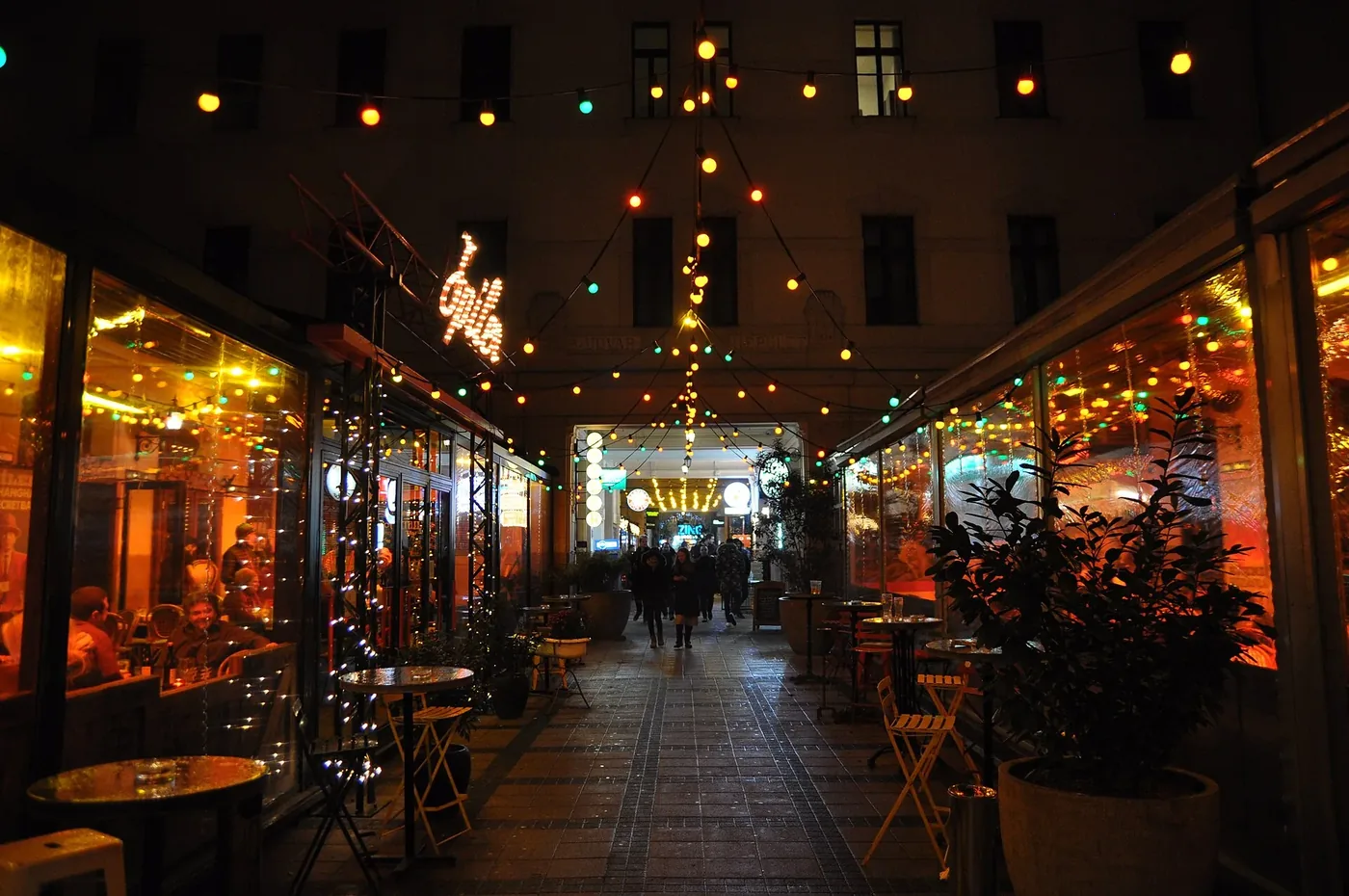 Gozsdu Udvar at night: glazed terraces glowing orange down both sides of the passage, strings of coloured bulbs strung overhead, bistro tables and folding chairs out on the paving, and a group of people walking toward a lit archway through to the next courtyard