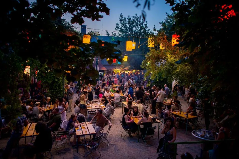 Dürer Kert's leafy open-air courtyard at dusk, paper lanterns strung over crowds at garden tables, Budapest