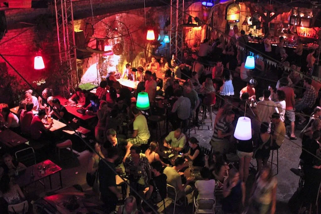 Szimpla Kert's packed open-air courtyard at night, lit by colourful glowing lamps under old brick arches — Budapest's original ruin bar