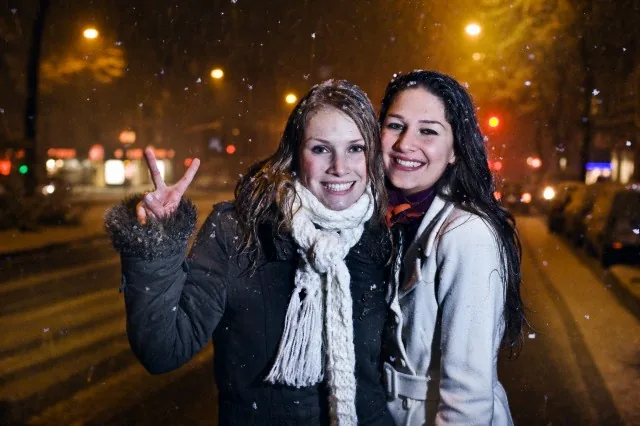 Two smiling guests in scarves and winter coats during falling snow on a Budapest pub crawl at night