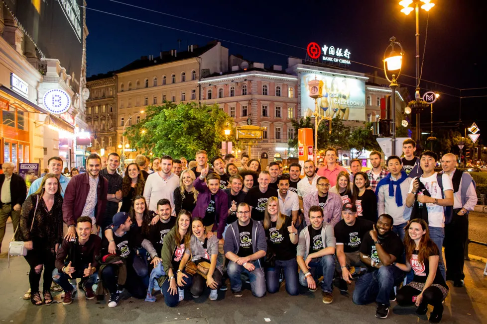A large group of pub crawl guests posed together on a floodlit Budapest boulevard at night, many wearing Pub Crawl Budapest T-shirts