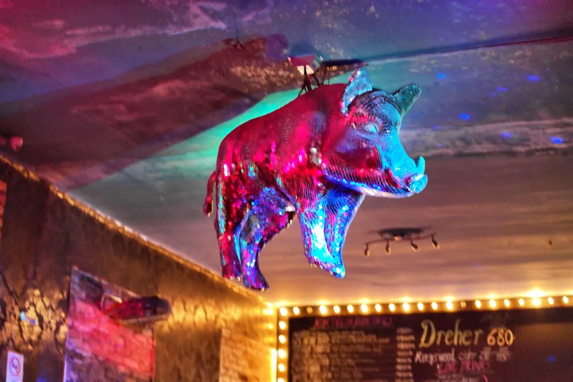 Looking up inside Instant-Fogas: a life-size pig covered in mirror-ball tiles hanging from the painted ceiling, lit magenta down one flank and cyan down the other by club spotlights, with a chalkboard drinks menu edged in warm filament bulbs on the wall below