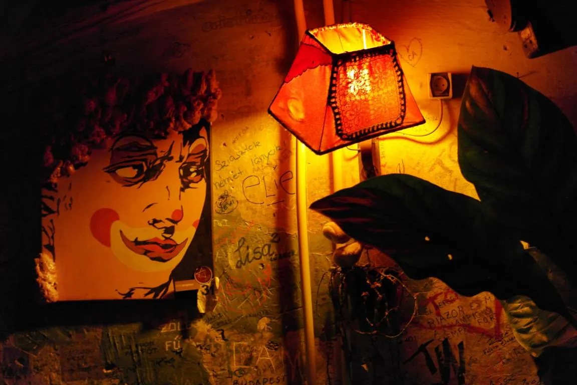 A dim corner inside Szimpla Kert at night: a red fringed lampshade glowing warm, a pop-art painting of a face on the wall beside it, and plaster covered in visitors' names and marker graffiti
