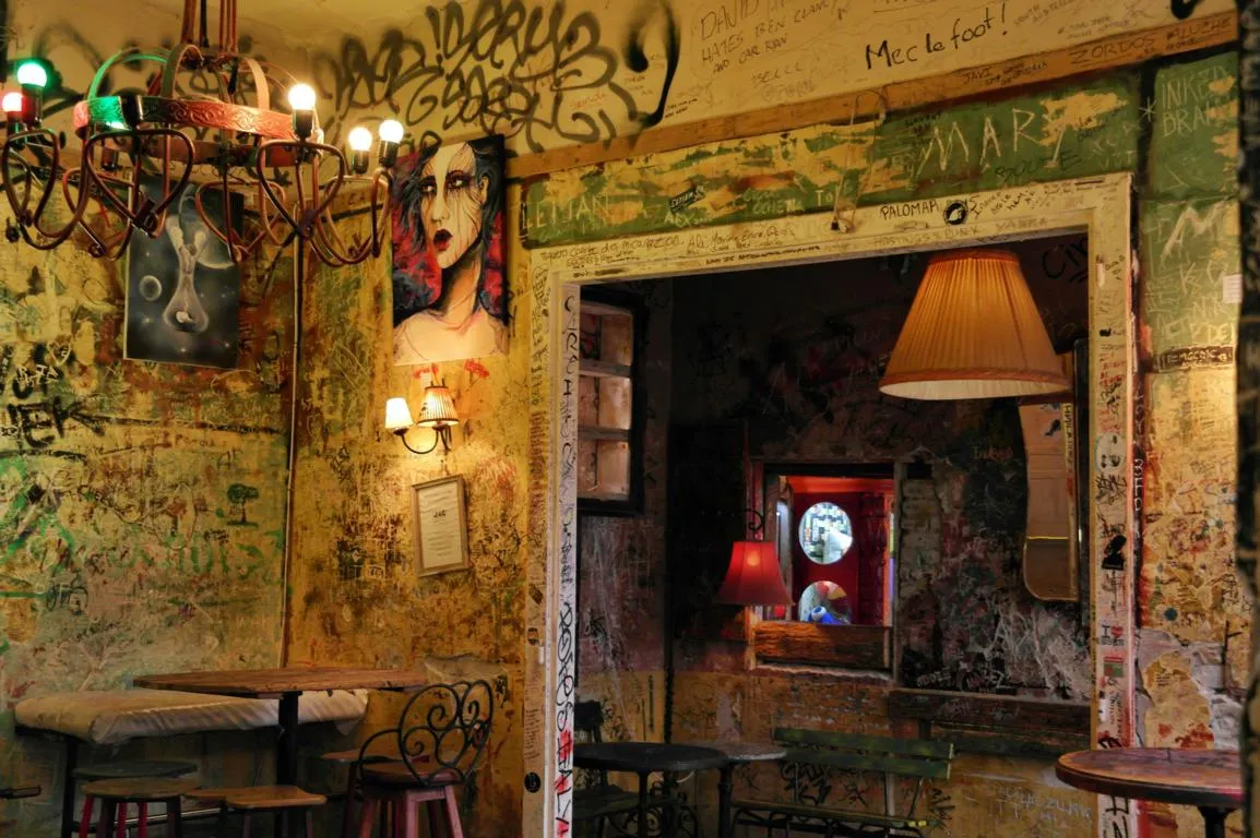 An empty room inside Szimpla Kert, every wall, beam and door frame covered in layered marker graffiti, a bare-bulb iron chandelier overhead, mismatched wooden tables and bar stools below, and a red-lit room visible through the far doorway