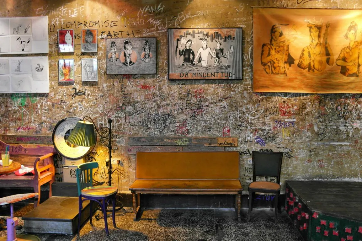 An empty room inside Szimpla Kert: a worn wooden bench and two mismatched chairs against a plaster wall scratched over with visitors' names and graffiti, a green pleated lamp lit beside a large round weighing scale, and framed drawings and a big ochre painting of three figures hung above