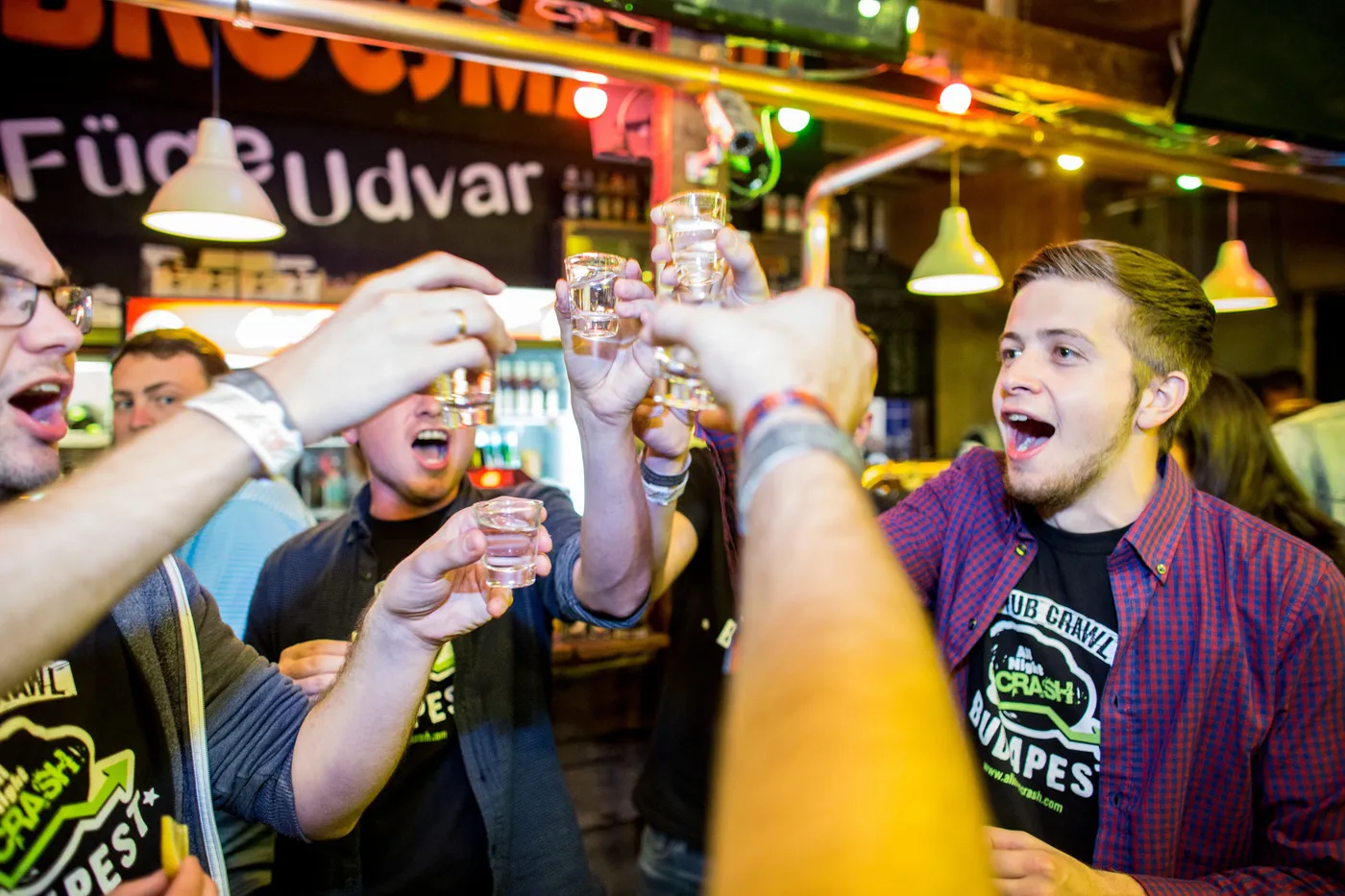 A group in black All Night Crash Pub Crawl Budapest T-shirts raising small glass shot measures in a toast at a bar counter, under a black sign reading Füge Udvar, with pendant lamps and a lit spirits shelf behind them