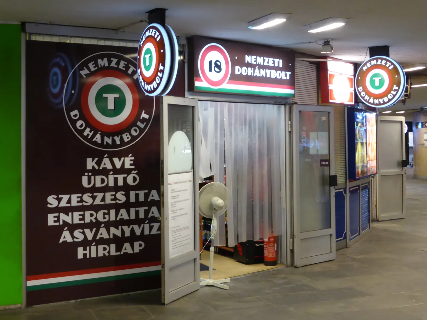 A Nemzeti Dohánybolt in a Budapest pedestrian underpass: illuminated circular signs carrying a green, white and red T logo, a red 18 age-restriction roundel, and a painted wall listing kávé, üdítő, szeszes ital, energiaital, ásványvíz and hírlap, with a plastic strip curtain hanging across the open doorway